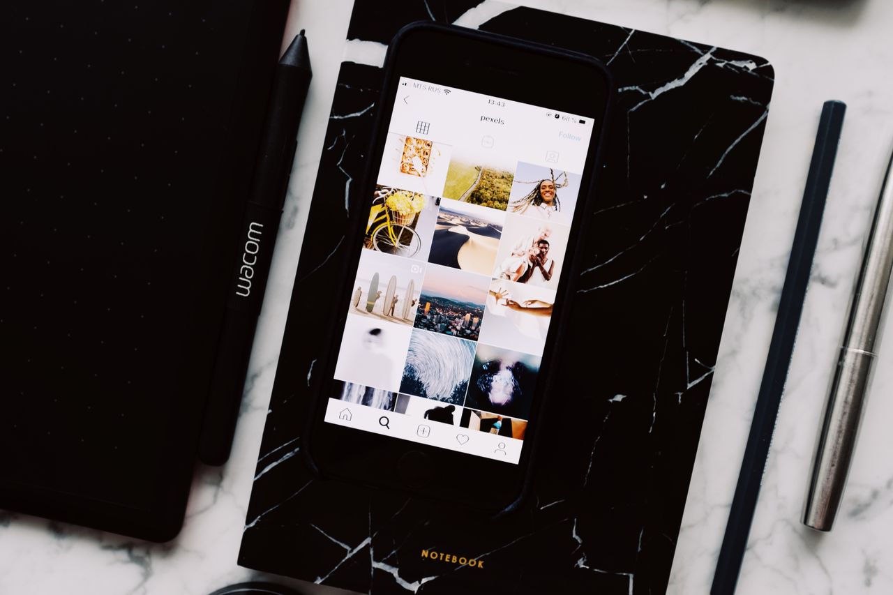 Top view of black notebook with cellphone above with pictures on screen and two pens aside located on marble table